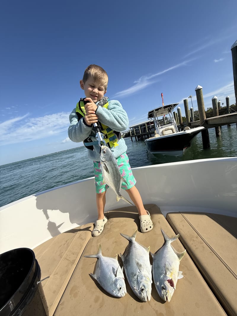 A young boy on a boat bow holding up a pompano with three more pompano on the deck, dock behind