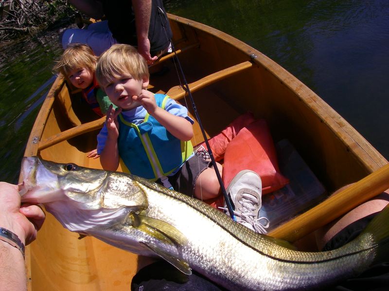 Two kids in a wooden paddle craft on a shaded creek with a snook held alongside the gunwale