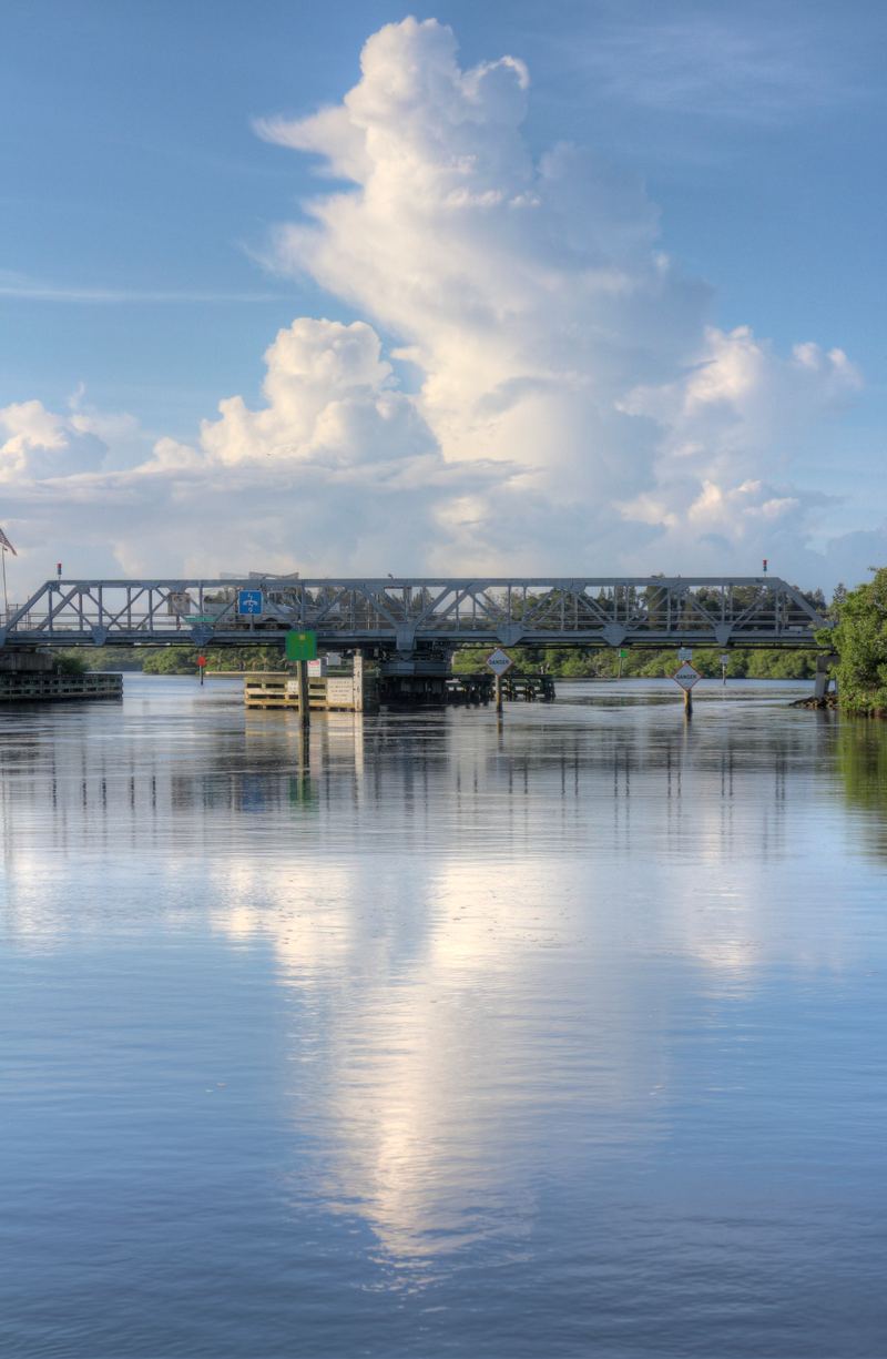 A low steel swing bridge over a narrow waterway, timber fender pilings at its base and clouds reflected in flat water