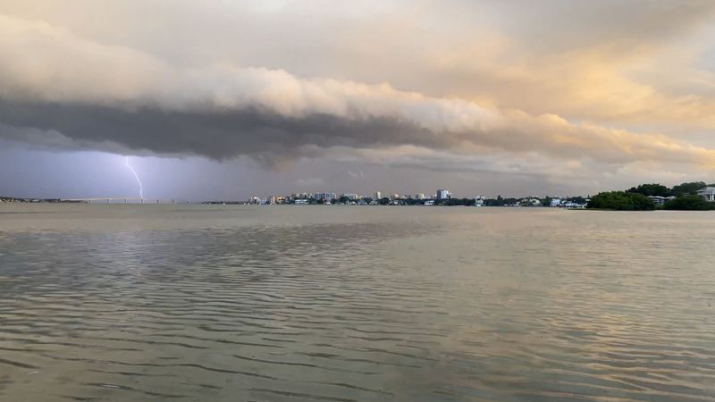 A lightning bolt drops out of a storm cell over open bay water, low shoreline in the distance