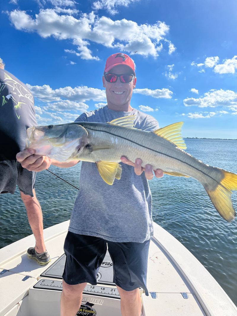 Two anglers on a boat bow, one holding a long snook horizontally over the gunwale, bay behind