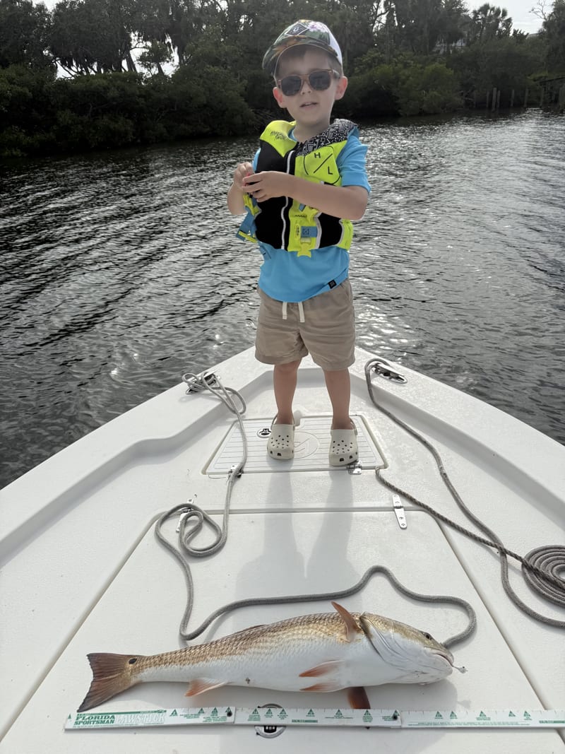 A young angler in a life jacket and sunglasses on the bow, his redfish laid on a measuring stick on the deck
