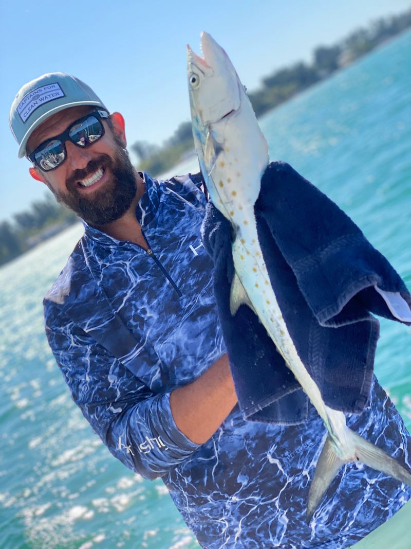 A Spanish mackerel held head-up over clear turquoise water, the yellow-gold flank spots that identify the species clearly visible