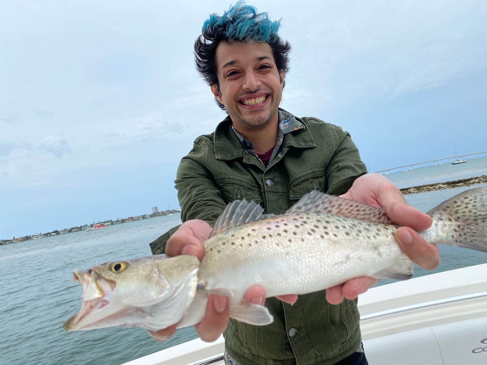 A grinning angler holding a heavy spotted seatrout on a bay boat, causeway bridge behind