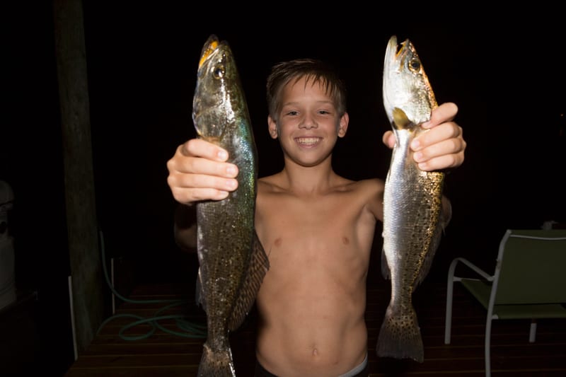 A young angler at night holding up two spotted seatrout, one in each hand, on a lit dock