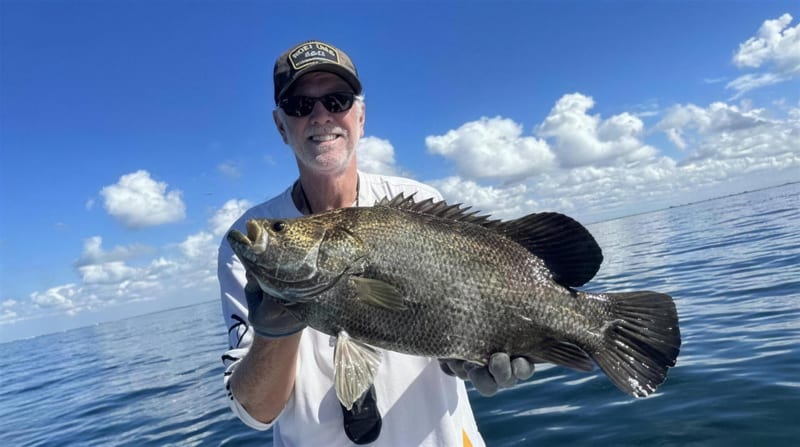 An angler holding a mottled tripletail over calm Gulf water under big cumulus clouds